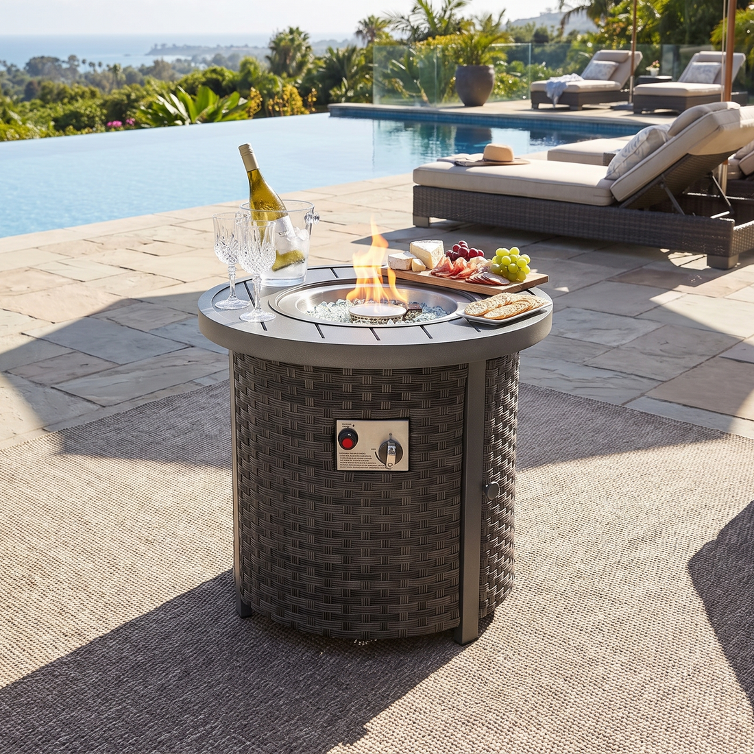 Compact round wicker fire pit table with snacks and a bottle on a stone patio beside a pool